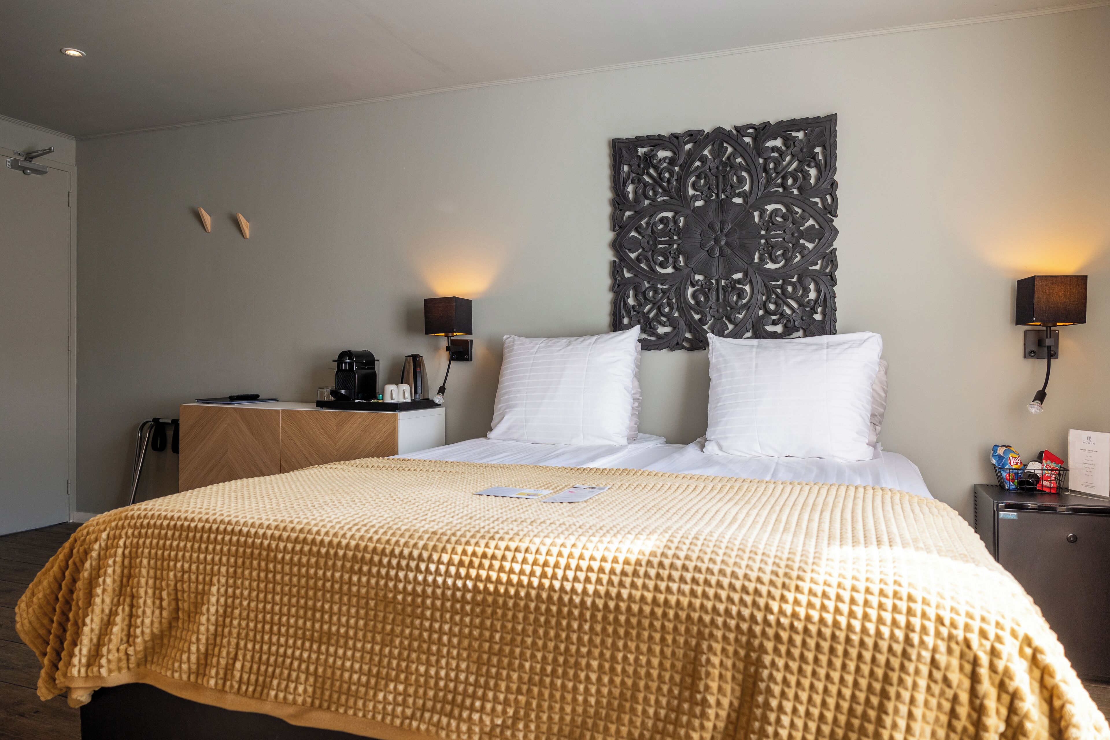 A hotel room with a large bed, a wooden nightstand, a decorative wall piece, and two wall-mounted lamps.