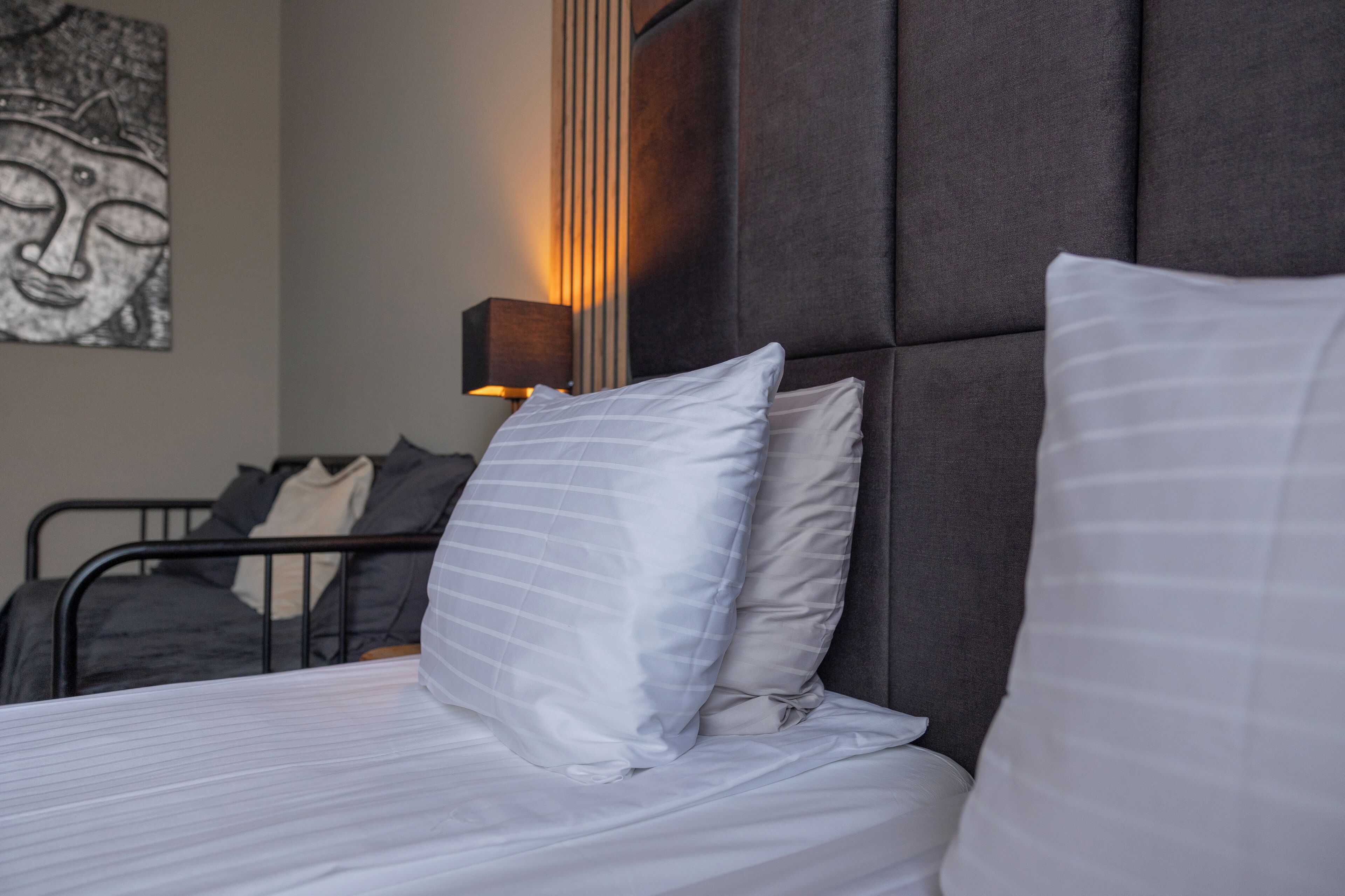 A neatly made bed with white linens and striped pillows, a dark upholstered headboard, and a wall-mounted lamp.
