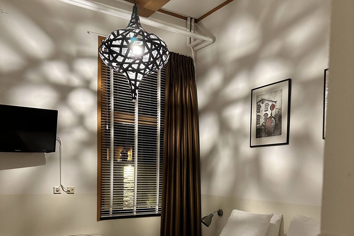 Un dormitorio con cama, televisor montado en la pared, radiador, ventana con persianas y una lámpara colgante.