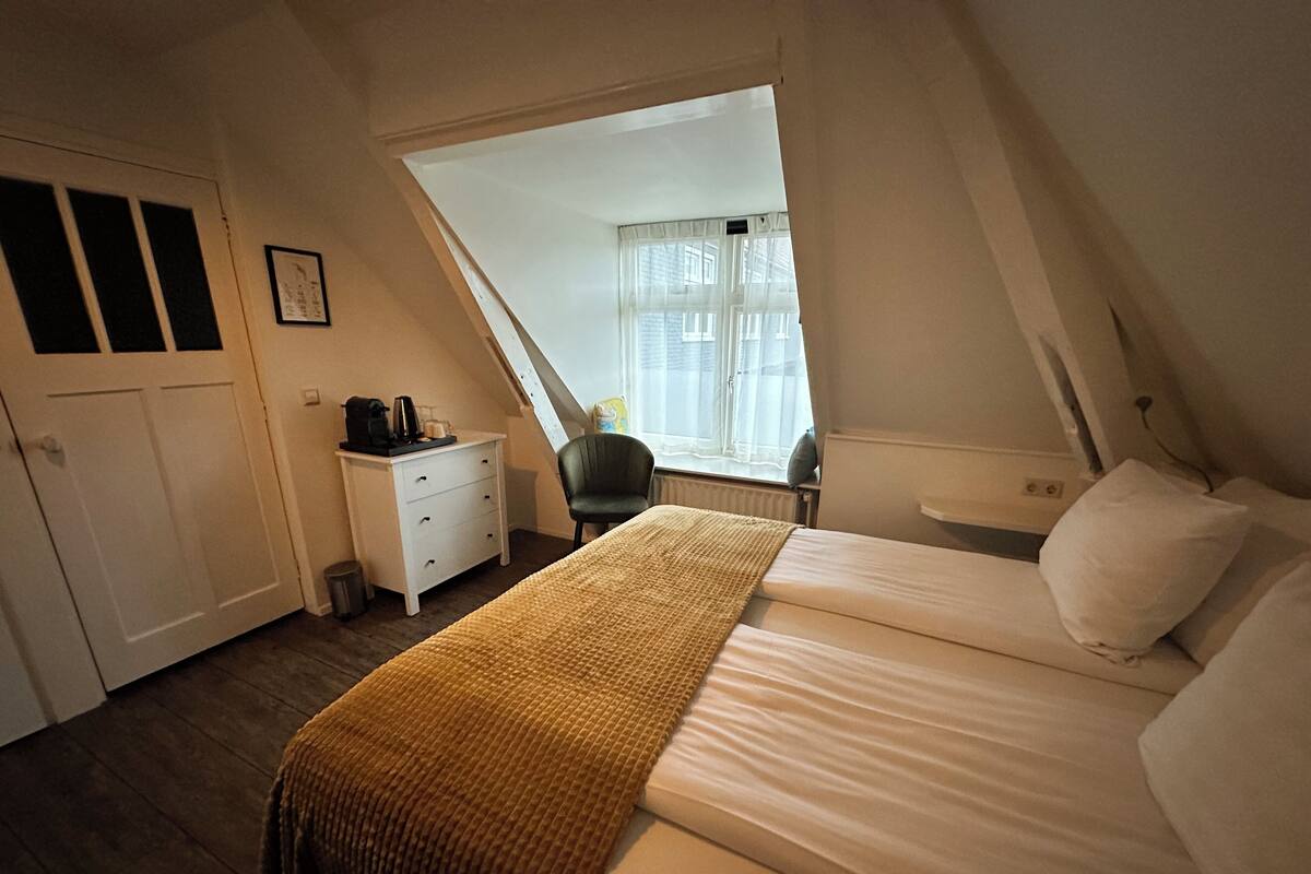 Un dormitorio con techo inclinado, una cama, una mesita de noche, una silla y una ventana con cortinas.