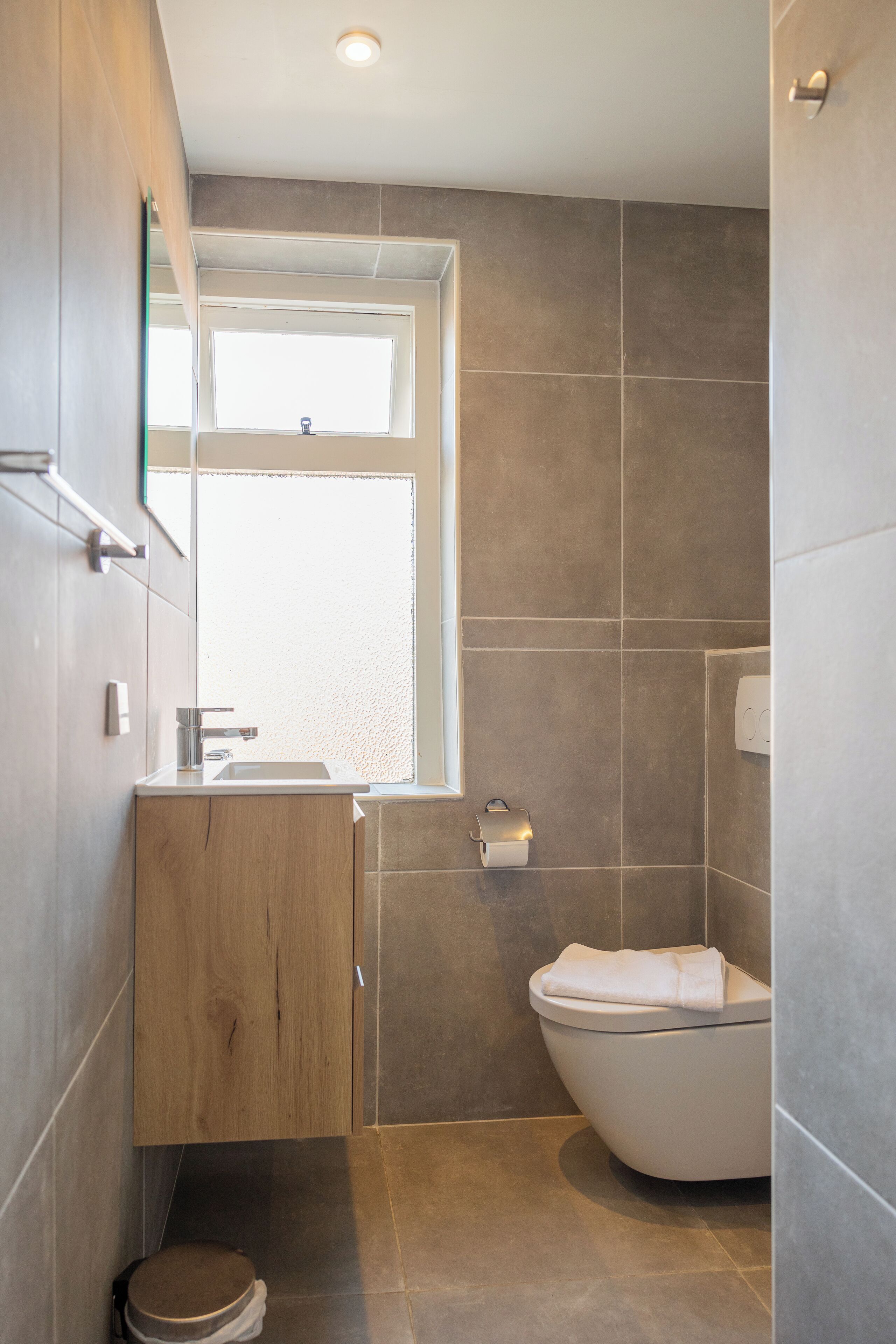 A modern bathroom with a wooden vanity, a toilet, and a window providing natural light.