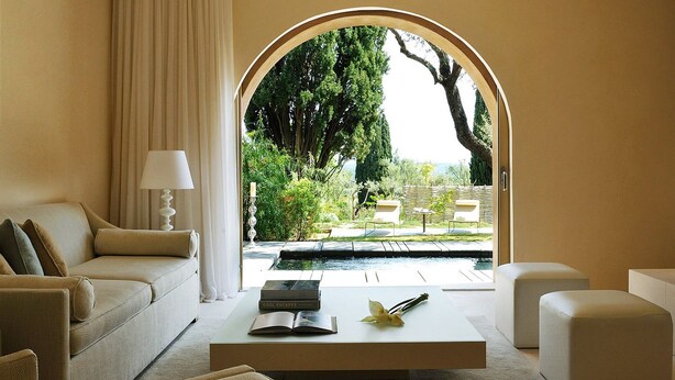 A modern living room with a sofa, armchairs, a coffee table, and a view of a garden through an arched doorway. - MUSE Saint Tropez (Ramatuelle)