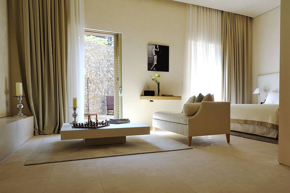 A modern hotel room with a large bed, a sofa, a small table with a chessboard, and a view of a stone wall outside.