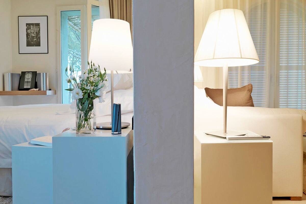A hotel room with a bed, a bedside table with a lamp, a small table with a vase of flowers, and a framed picture on the wall.