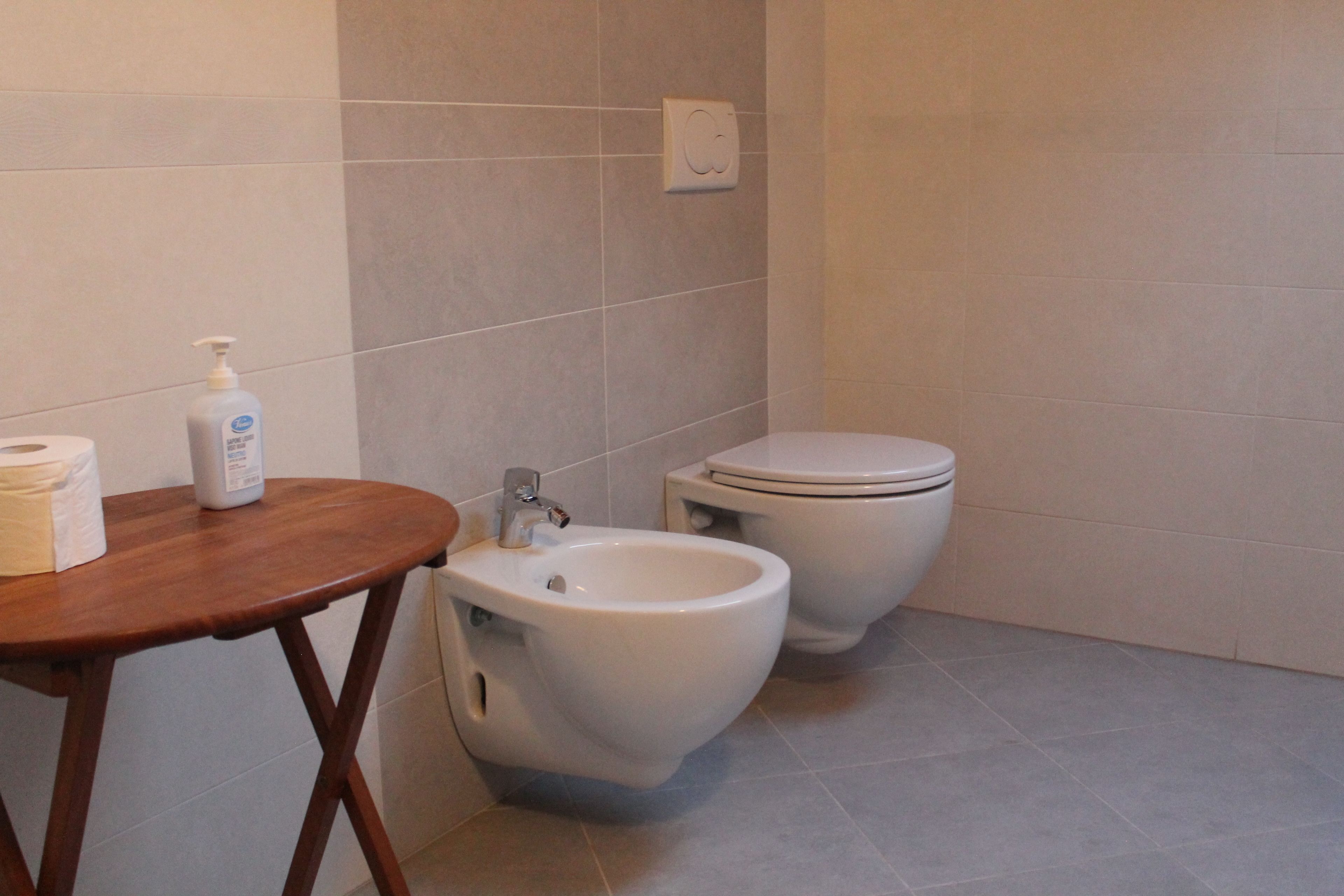 Un baño con bidé, inodoro y una mesa de madera con una botella de jabón y papel higiénico.
