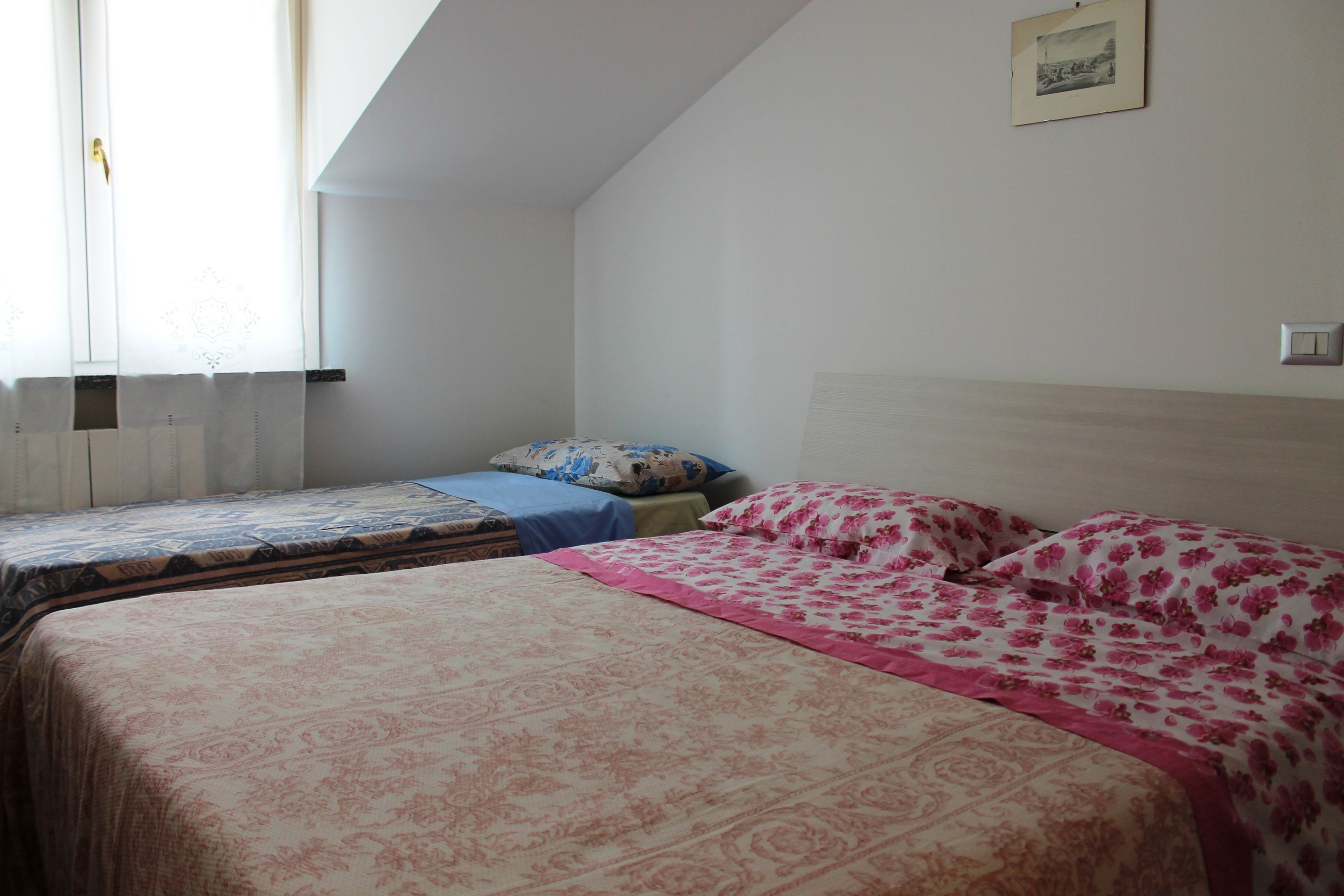 Ein Schlafzimmer mit zwei Betten, einem Fenster mit Vorhängen, einem Bild an der Wand und einem Lichtschalter.