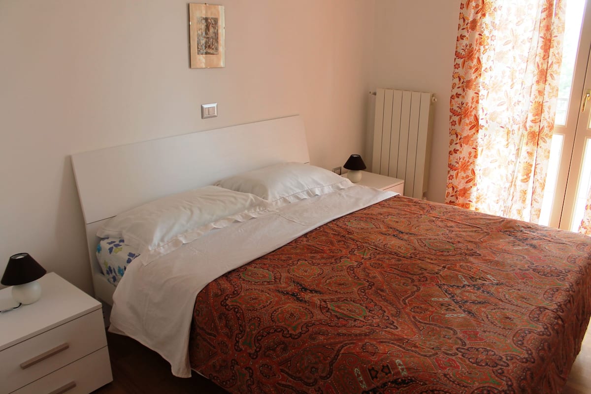 A bedroom with a bed, a nightstand, a lamp, a radiator, and a window with curtains.
