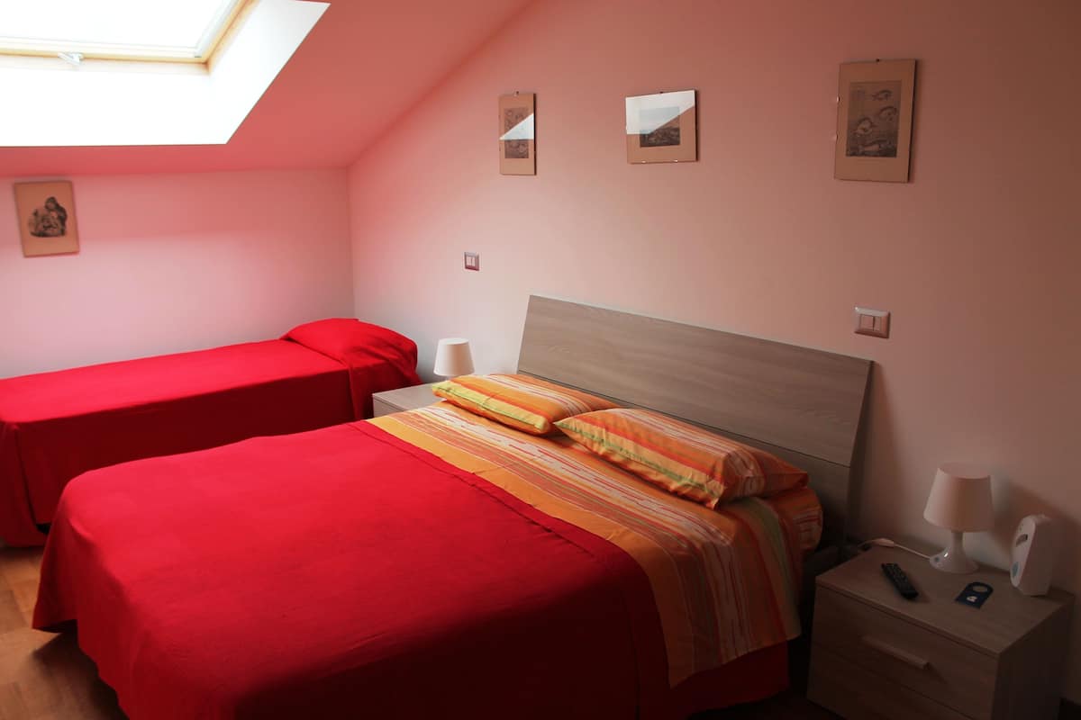 A bedroom with two beds, one with a red bedspread and the other with a striped bedspread. There is a nightstand with a lamp and a remote control.