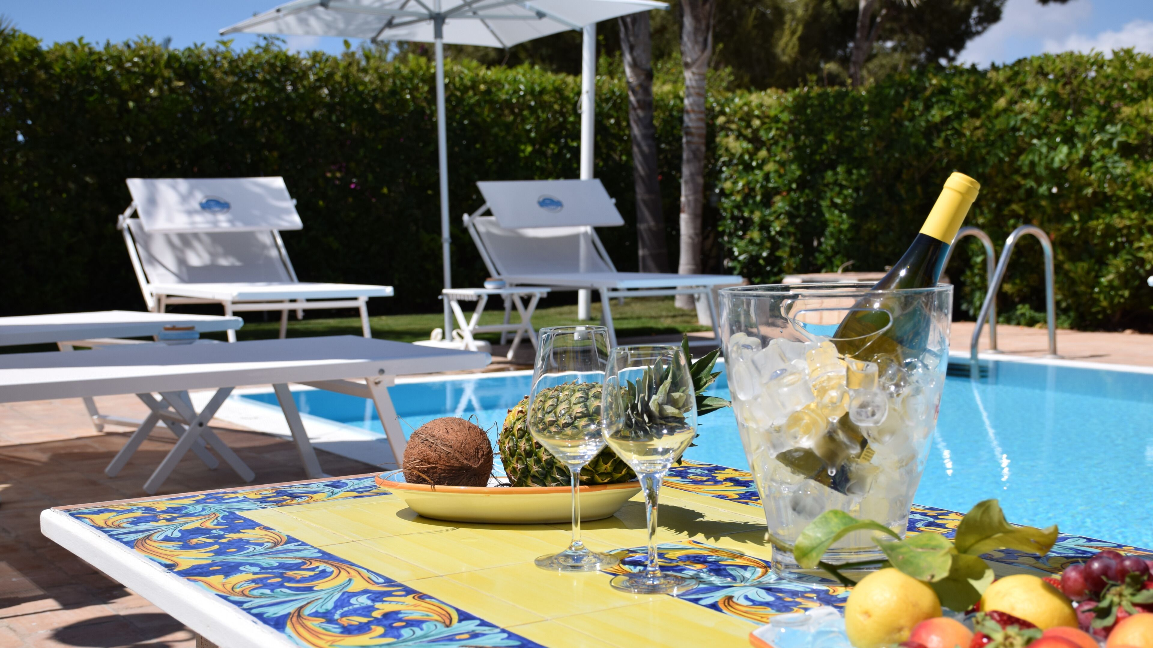 Un tavolo a bordo piscina con un'elegante composizione di frutta tropicale, una bottiglia di champagne e calici di vino.
