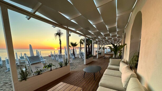 Un salón junto a la playa con suelo de madera, asientos acolchados y vistas a la puesta de sol sobre el océano.