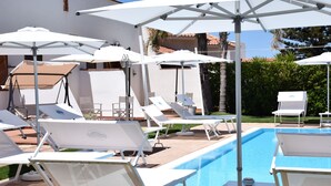 Zona junto a la piscina con múltiples tumbonas y sombrillas, una piscina azul cristalina y un edificio blanco al fondo.