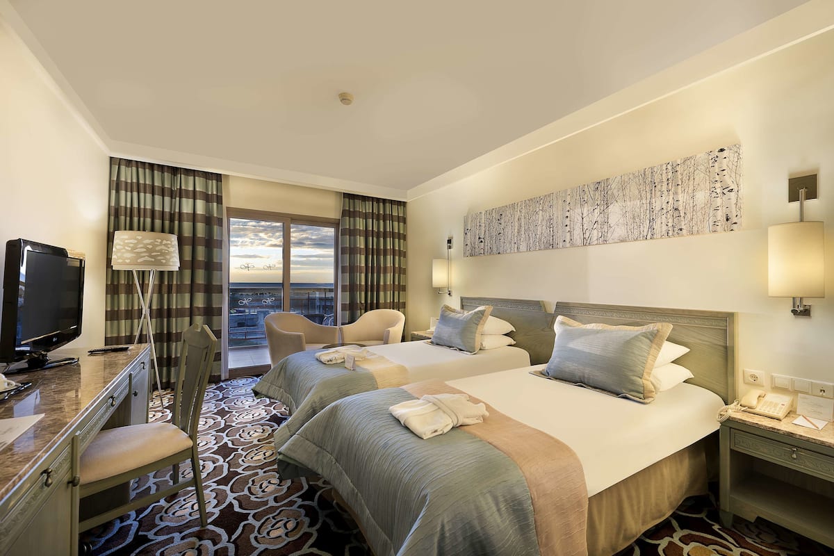 A hotel room with two beds, a desk, a TV, and a balcony with curtains.
