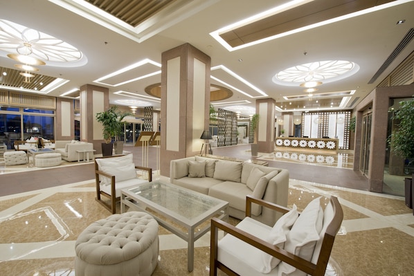 Un vestíbulo amplio con diseño moderno, que cuenta con una zona de estar central con sofá y reposapiés, una mesa de centro de vidrio y una pared decorativa con estampado floral.