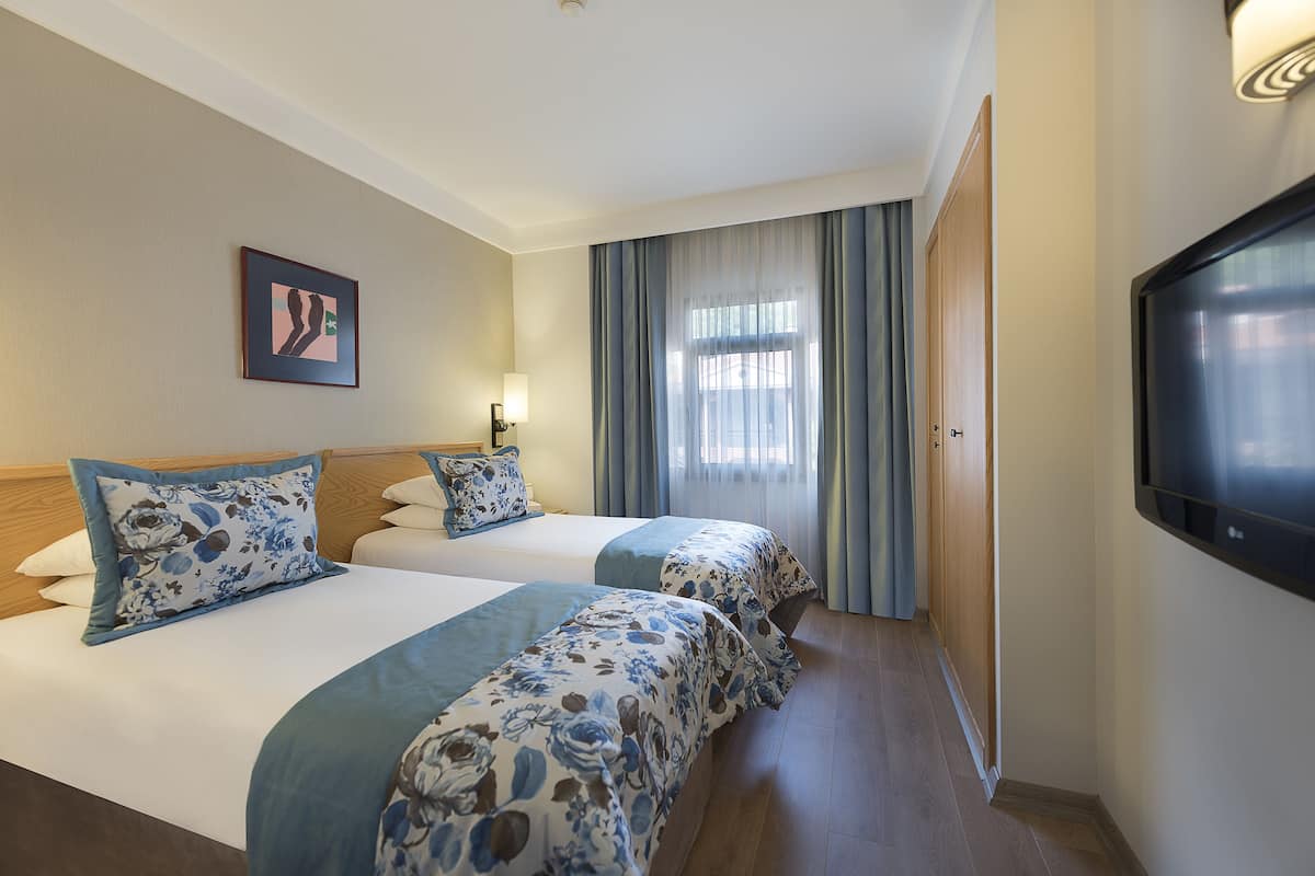 A hotel room with two beds, a TV, a window with curtains, and a framed picture on the wall.