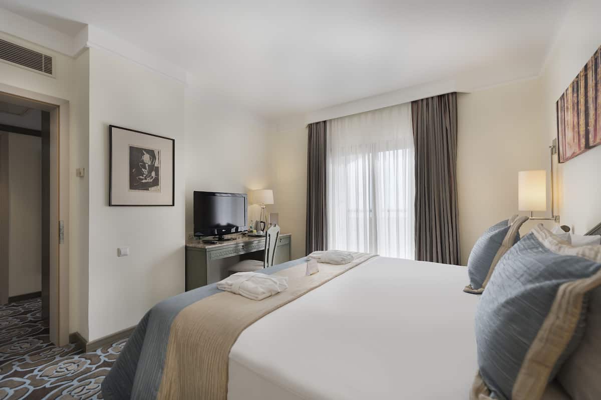 A hotel room with a large bed, a desk with a computer, a television, and a window with curtains.