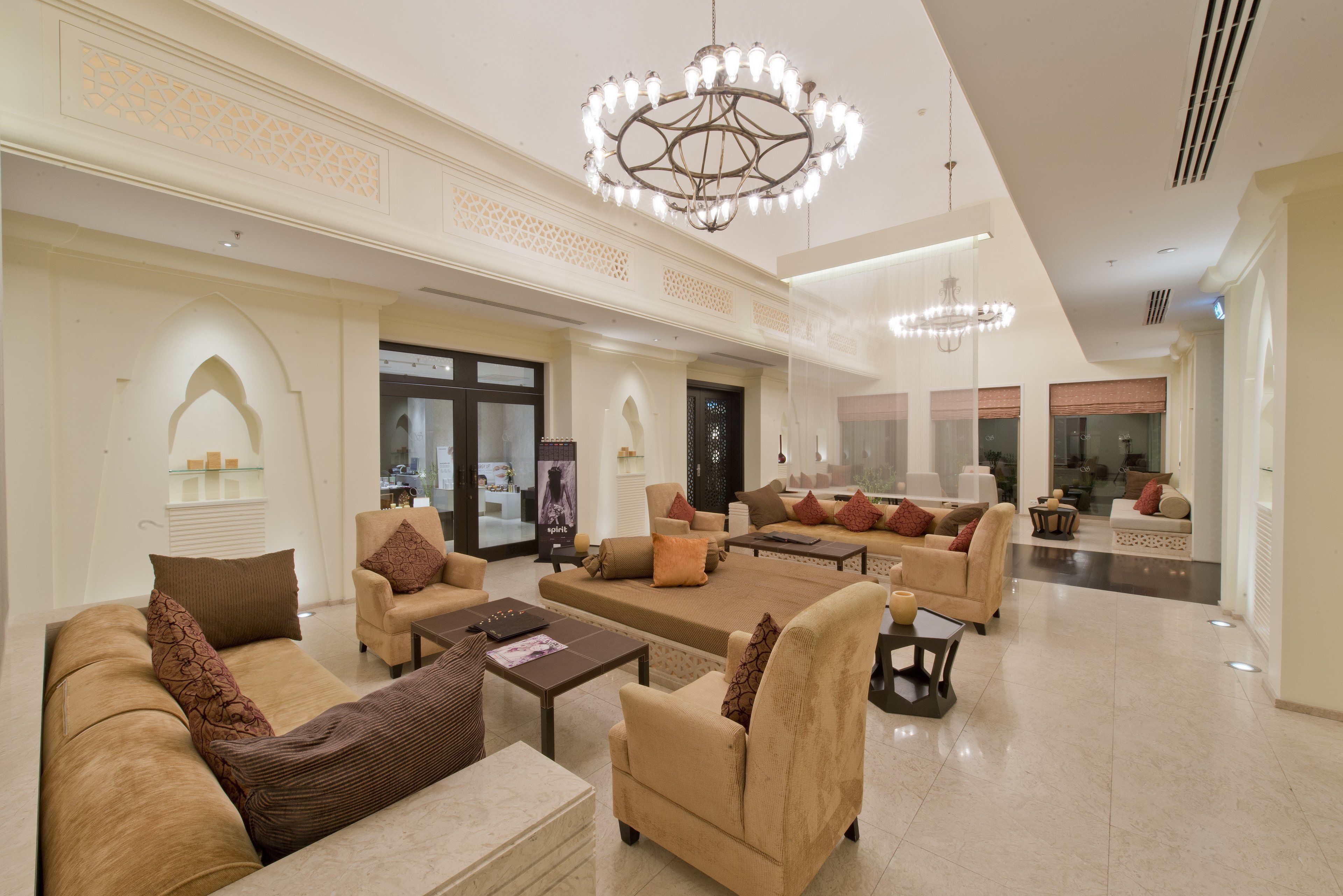A spacious lobby with multiple seating areas, including sofas and armchairs, arranged around a central coffee table. There are decorative chandeliers hanging from the ceiling and a large glass door leading to another room.