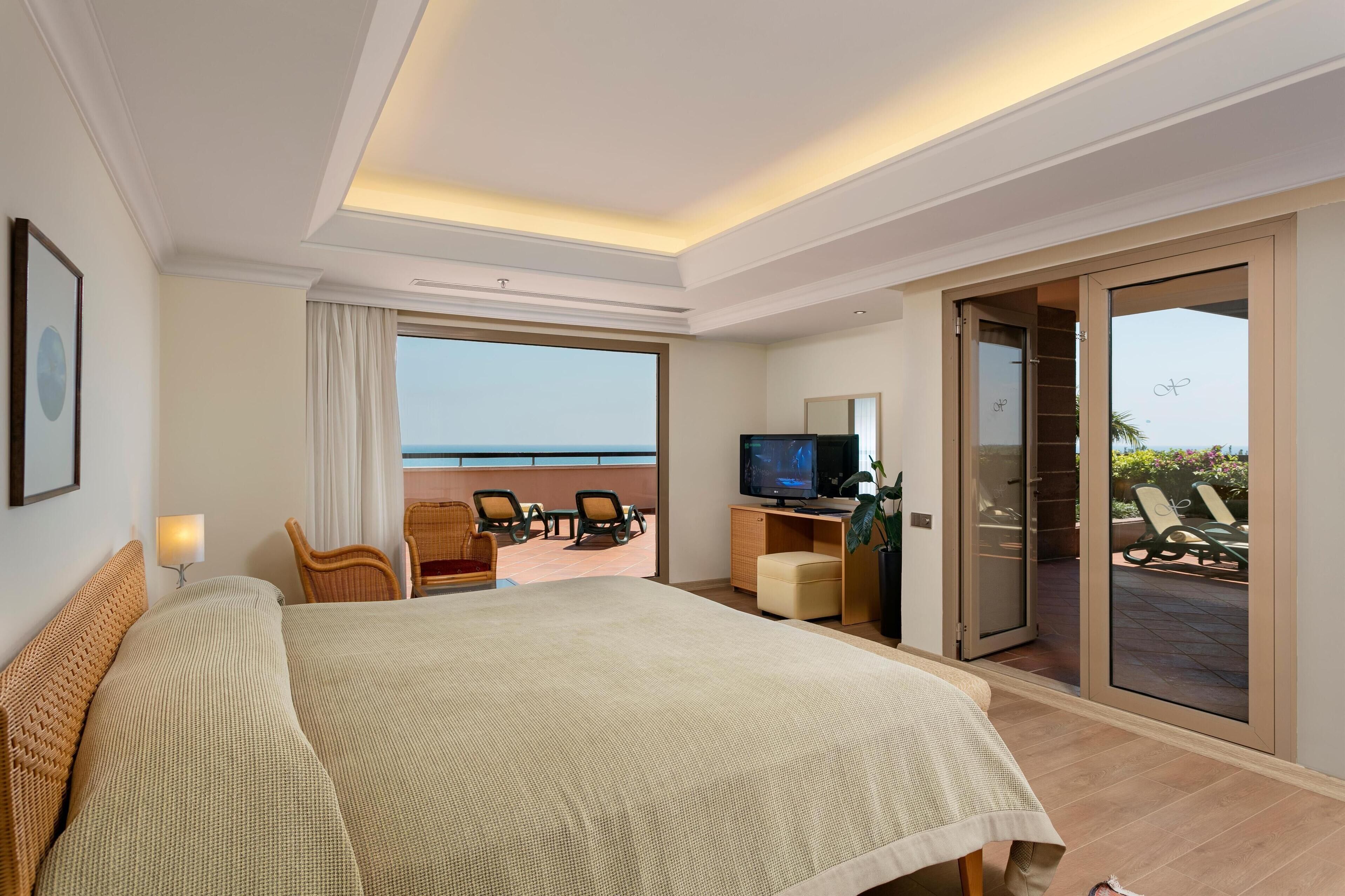 A hotel room with a large bed, a TV, a desk, and a balcony with a view of the sea.