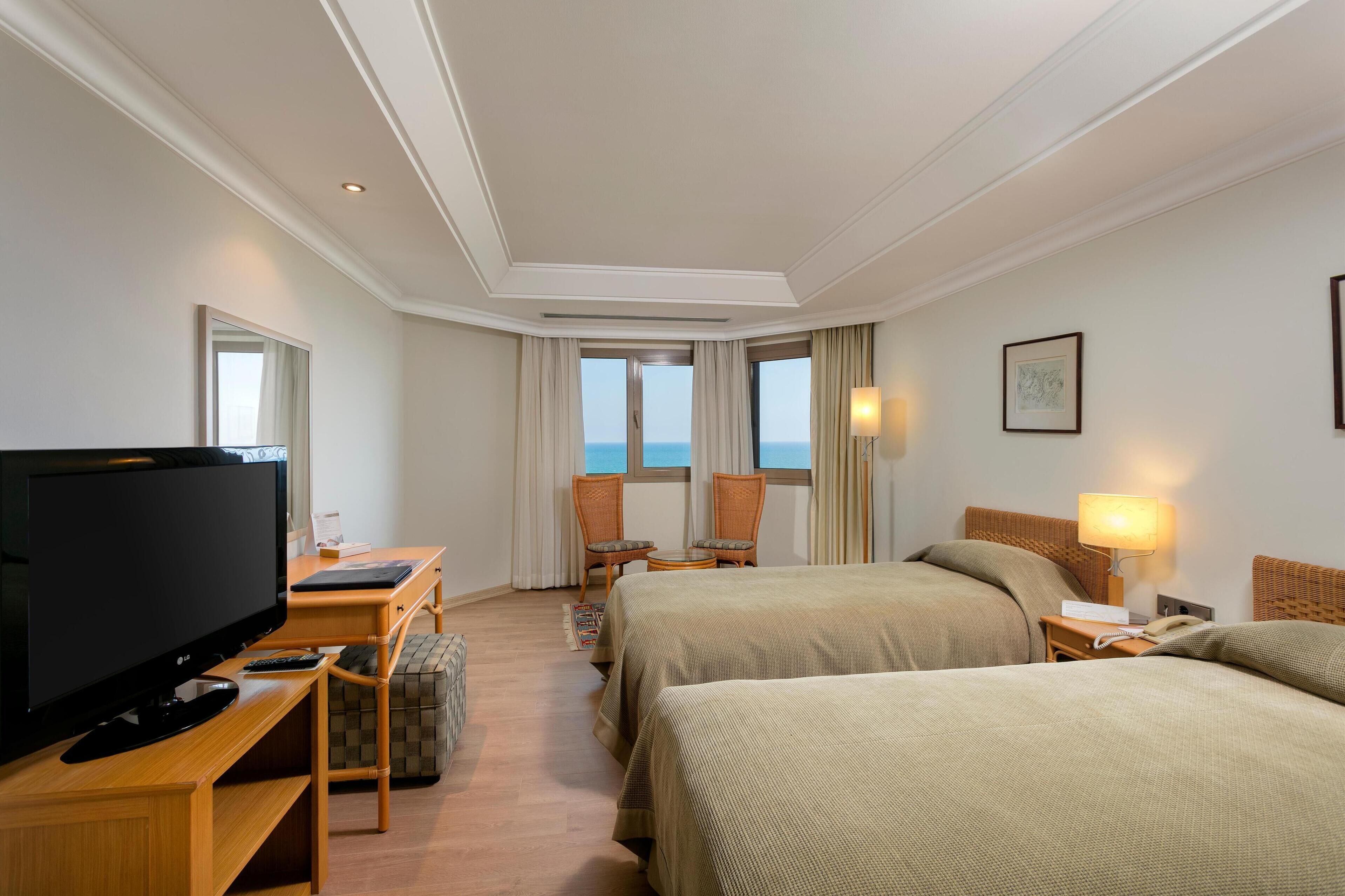 Een hotelkamer met een televisie, twee bedden, een bureau, een stoel, een lamp en uitzicht op zee.
