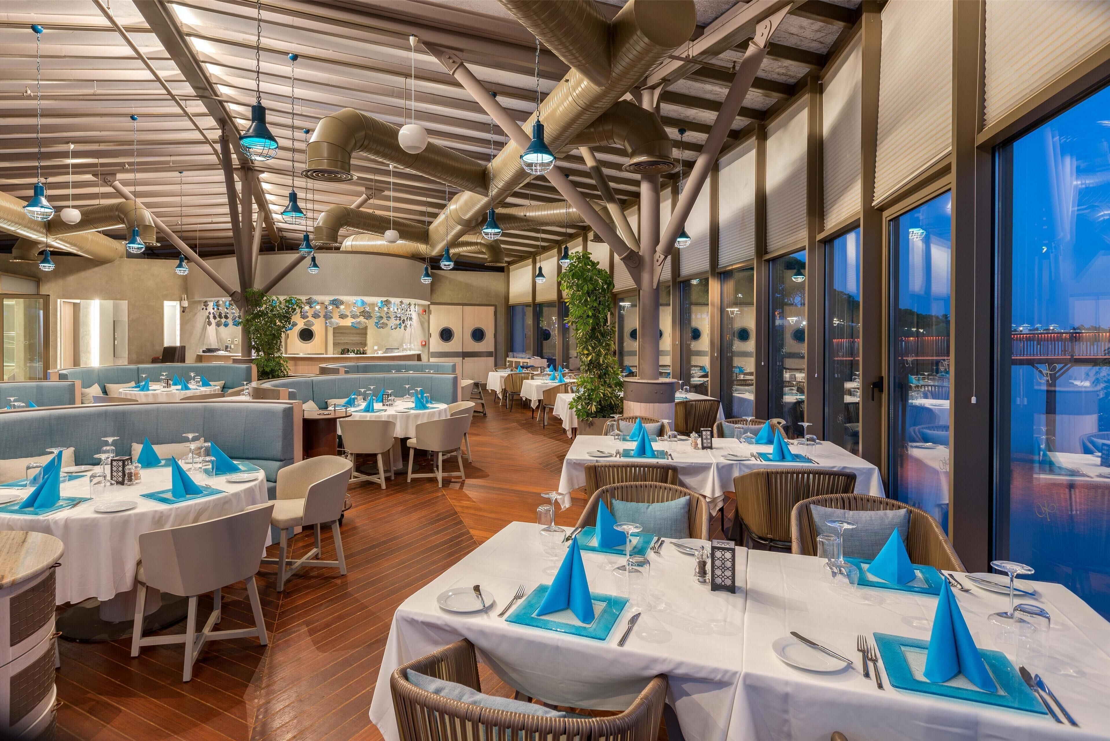 Ein Restaurant mit hohen Decken, freiliegenden Lüftungsrohren und großen Fenstern mit Blick auf die Stadt. Die Tische sind mit blauen Servietten und weißen Tischdecken gedeckt.