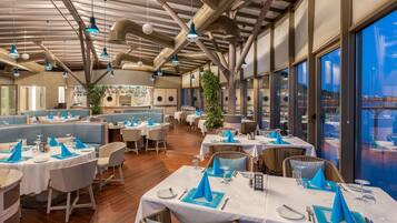 Ein Restaurant mit hohen Decken, freiliegenden Lüftungsrohren und großen Fenstern mit Blick auf die Stadt. Die Tische sind mit blauen Servietten und weißen Tischdecken gedeckt.
