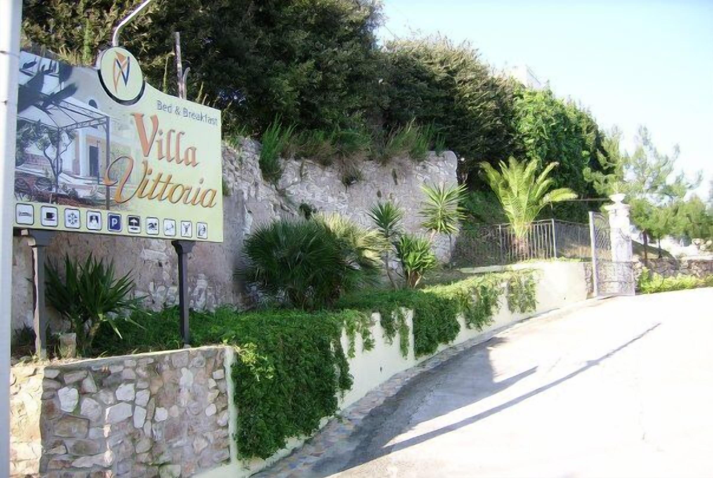 Une enseigne pour la Villa Vittoria, avec un symbole représentant un hébergement de type chambre d’hôtes, un mur en pierre et une végétation luxuriante.
