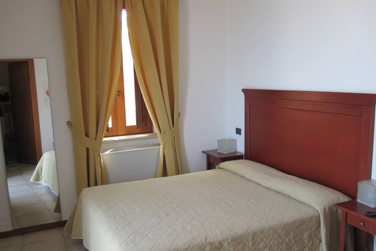 A bedroom with a bed, a wooden headboard, a nightstand, a window with curtains, and a mirror.