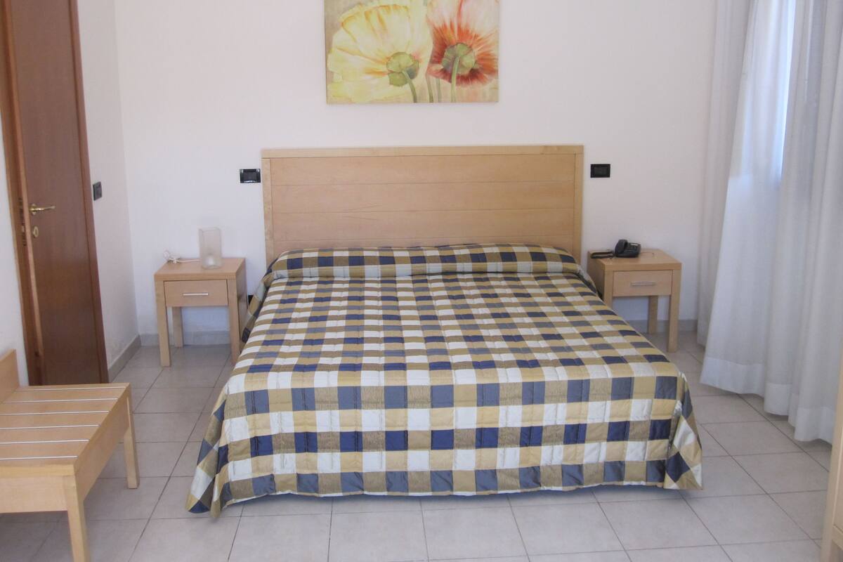A bedroom with a checkered bedspread, a wooden headboard, two bedside tables, a nightstand, a wooden chair, and a painting on the wall.