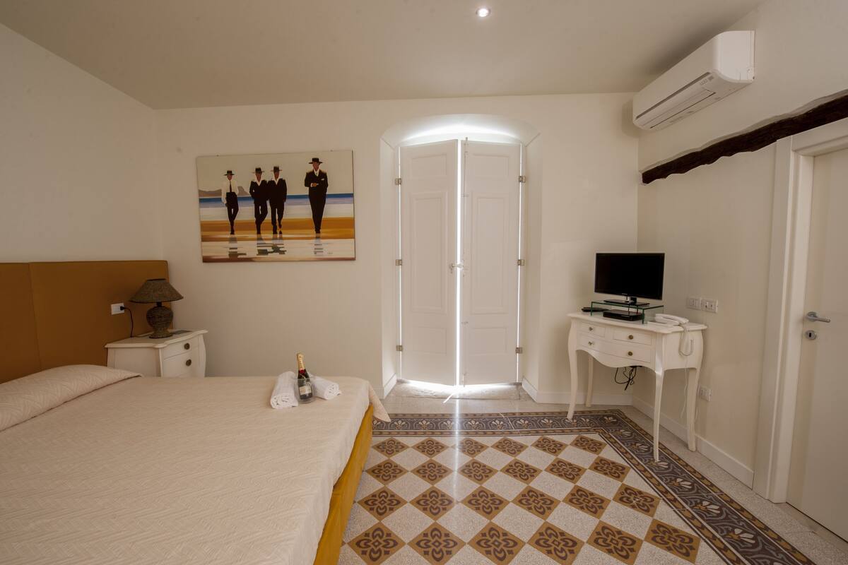 Habitación de hotel con cama, escritorio con televisor, un cuadro en la pared y una puerta con marco iluminado.