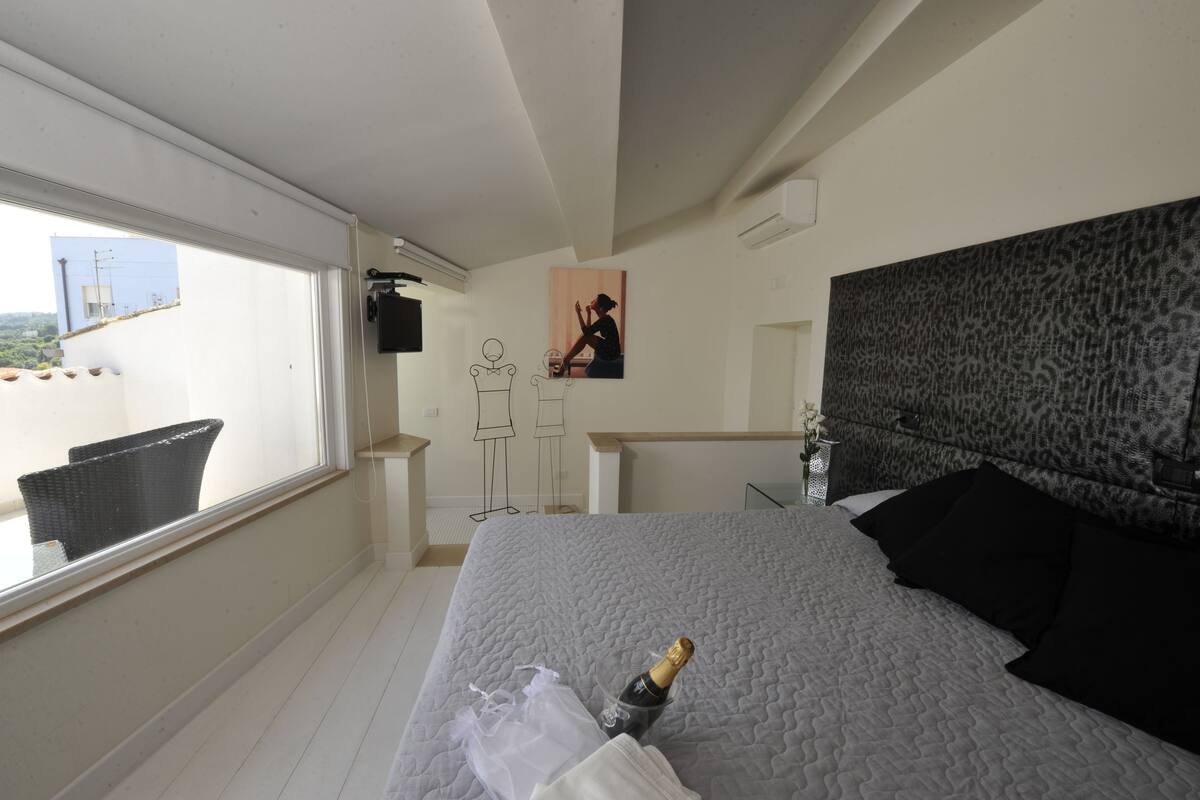 Habitación de hotel con cama, ventana, balcón, silla, escritorio y un cuadro en la pared.