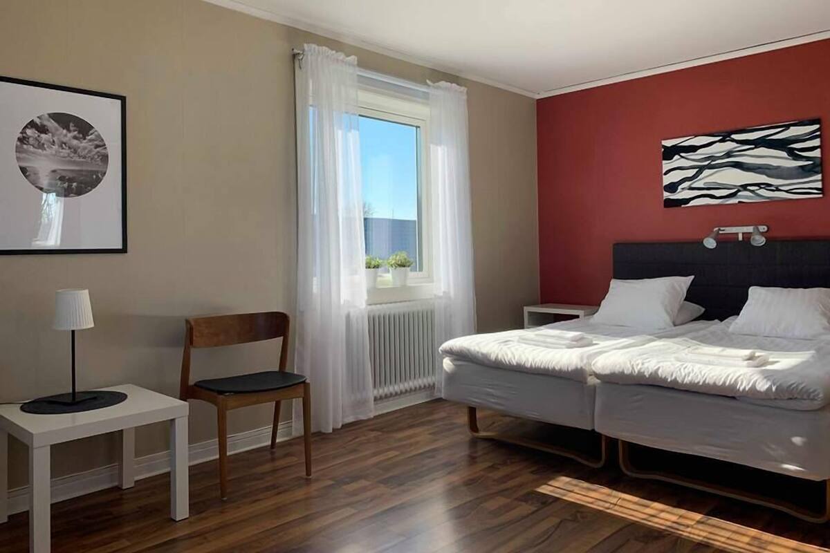 A hotel room with a bed, a chair, a small table, a window with curtains, and a wall with a framed picture.