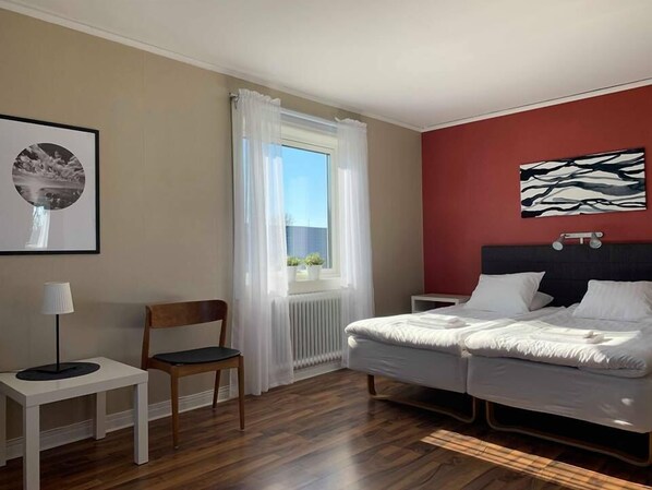 A hotel room with a bed, a chair, a small table, a window with curtains, and a wall with a framed picture. - Gamla Bryggeriets B&B (Kopingsvik)