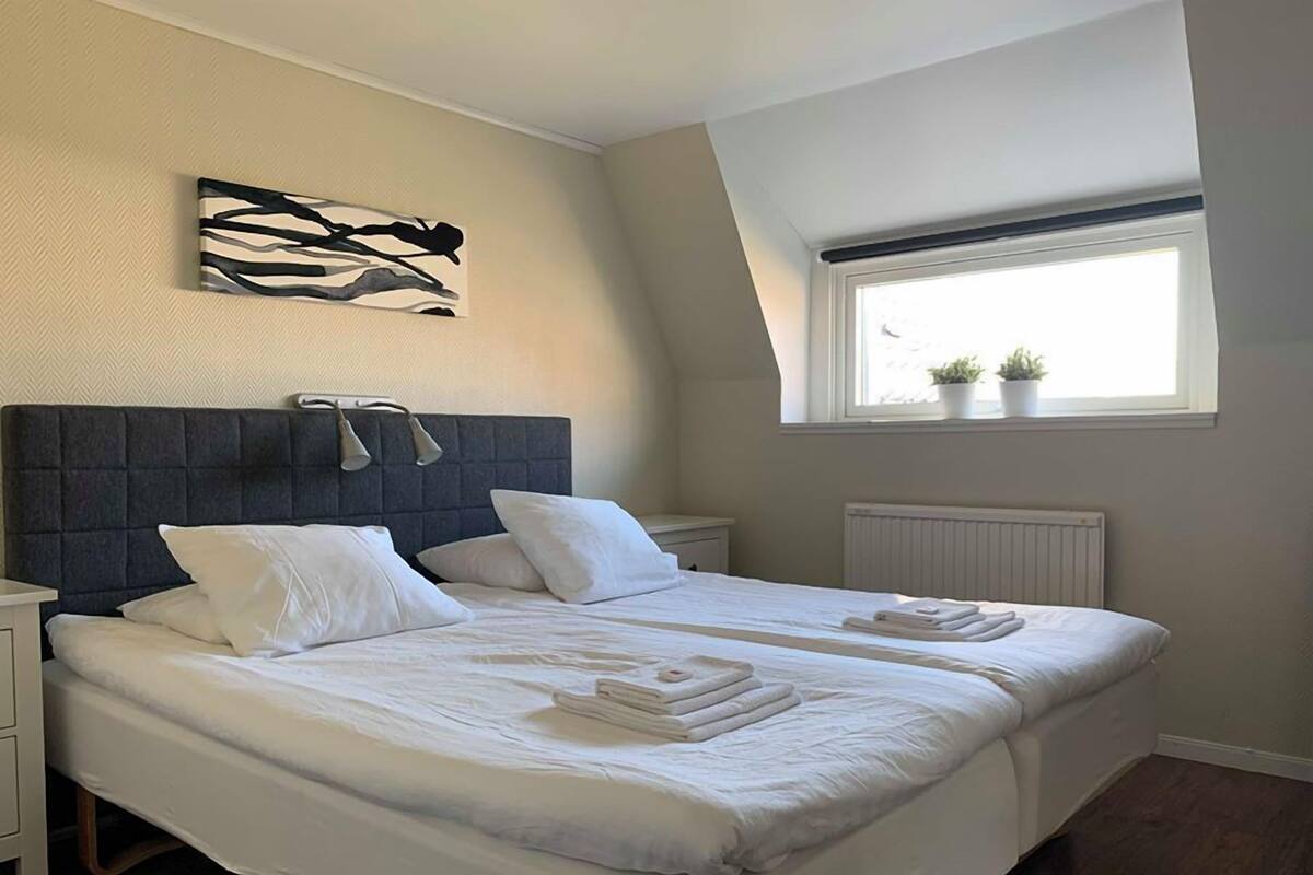 A bedroom with a large bed, a headboard, a nightstand, a window with plants, and a wall art piece.