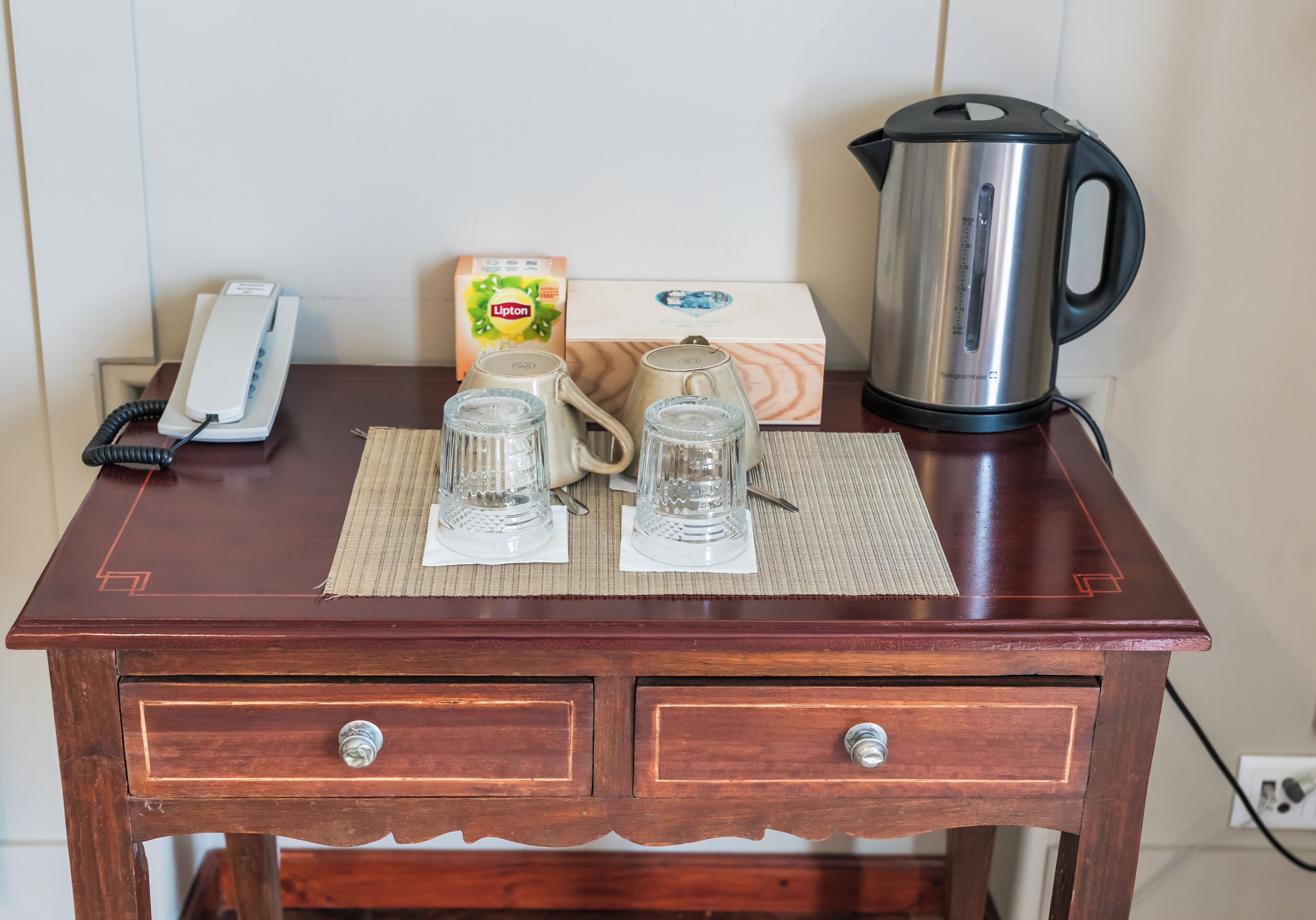 A wooden table with a telephone, Lipton tea box, two glass mugs, a kettle, and a box of tea bags.