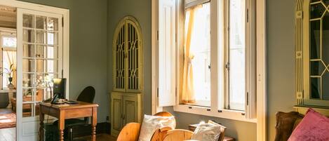Une pièce avec un bureau en bois, une chaise en bois, un canapé avec des coussins et une grande fenêtre avec des rideaux.