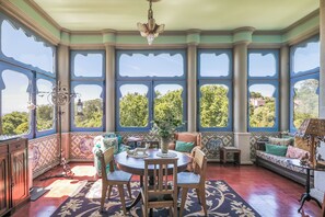 A room with a large bay window, a dining table with chairs, a sofa, and a small table with a lamp. - Chalet Saudade (Sintra)