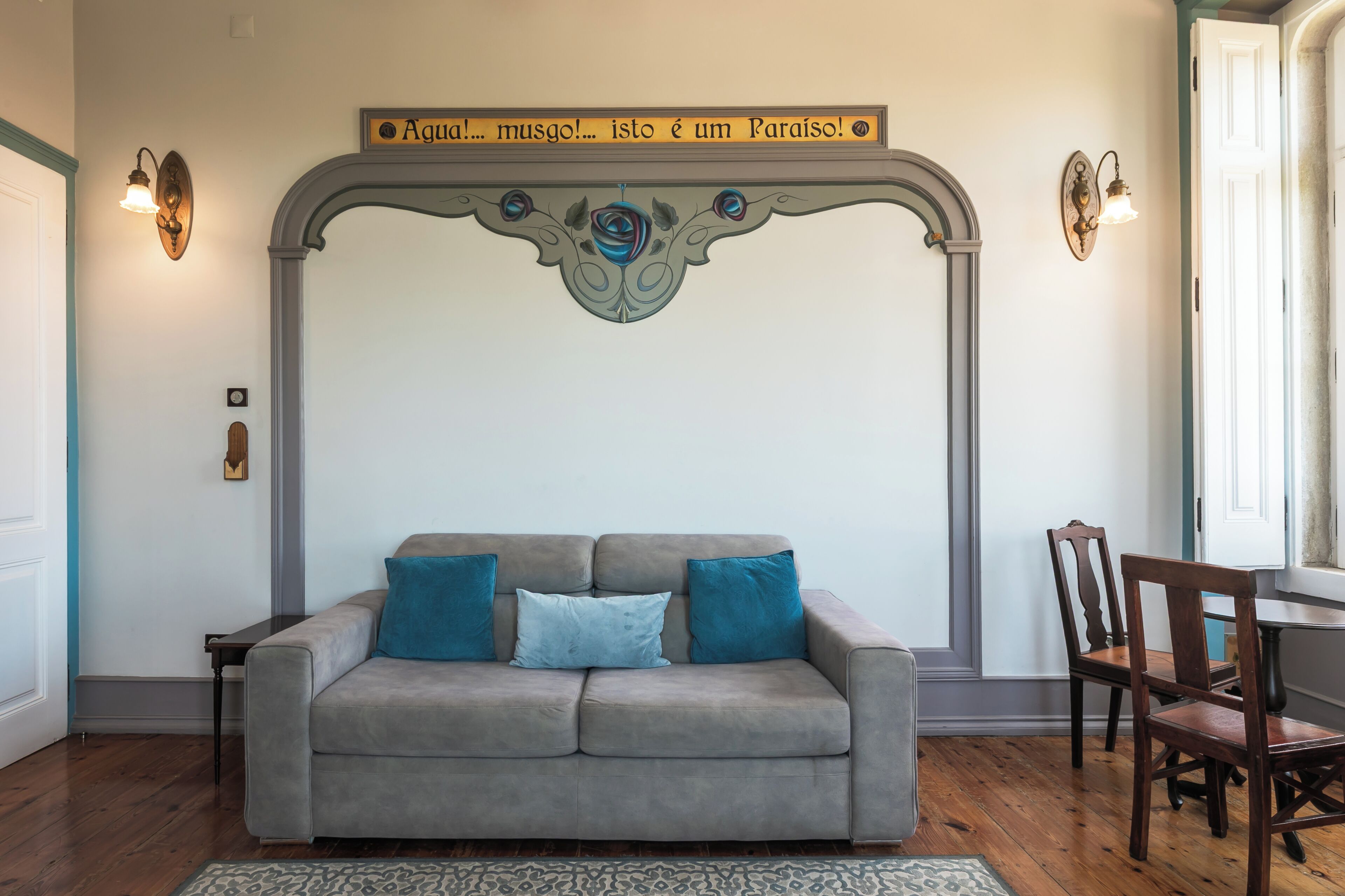 A living room with a grey sofa, two blue cushions, a patterned rug, a wooden chair, and a decorative wall with a sign.