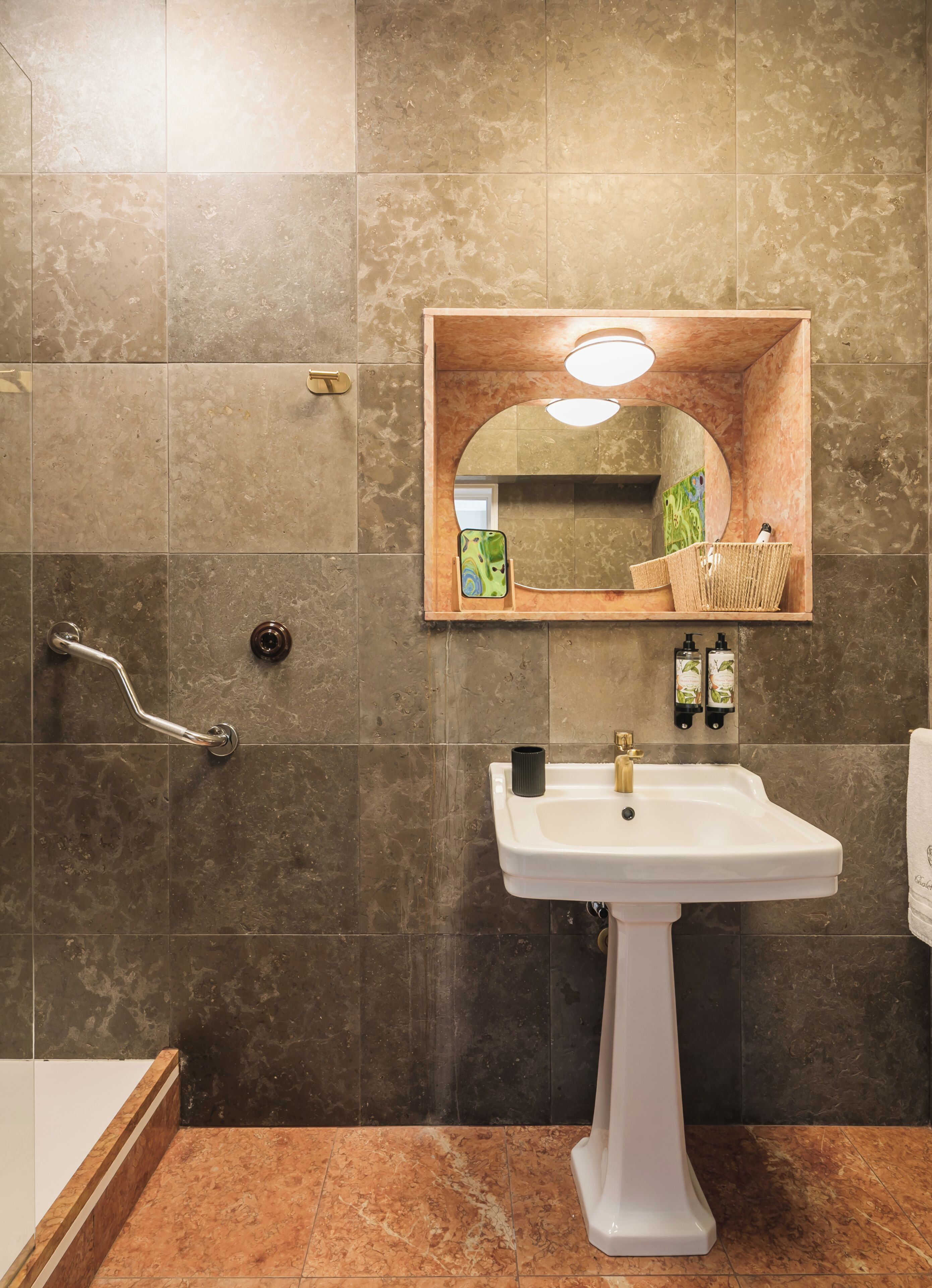 A bathroom with a white pedestal sink, a mirror with a wooden frame, and a handrail.