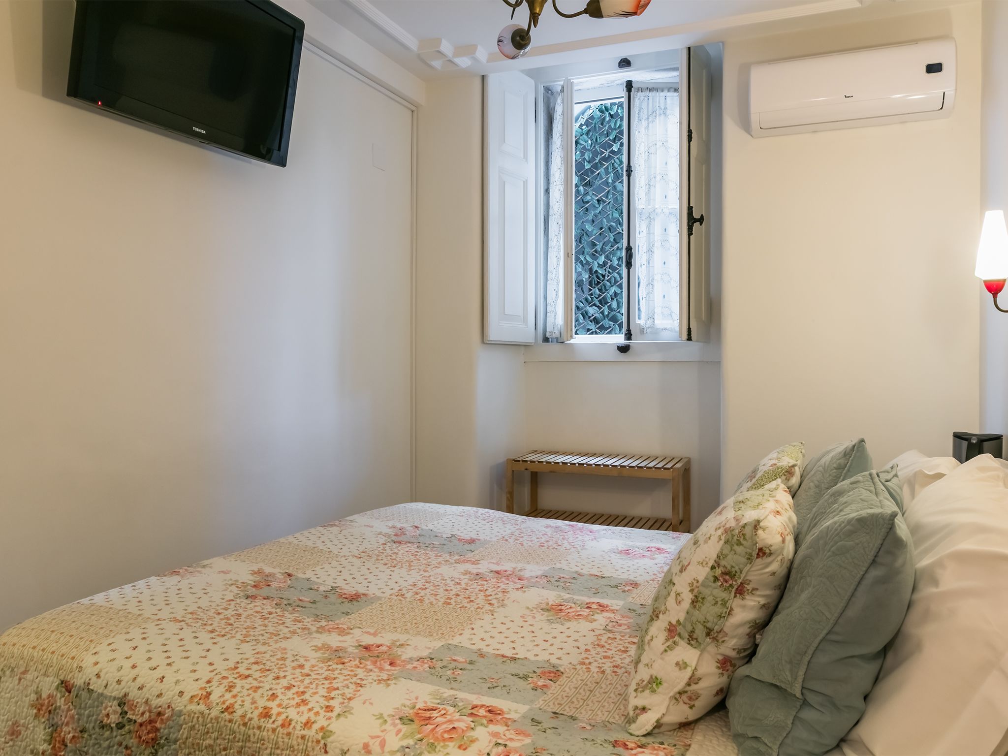 Ein Schlafzimmer mit einem Bett, einem an der Wand montierten Fernseher, einem Fenster mit Vorhängen und einem Nachttisch.