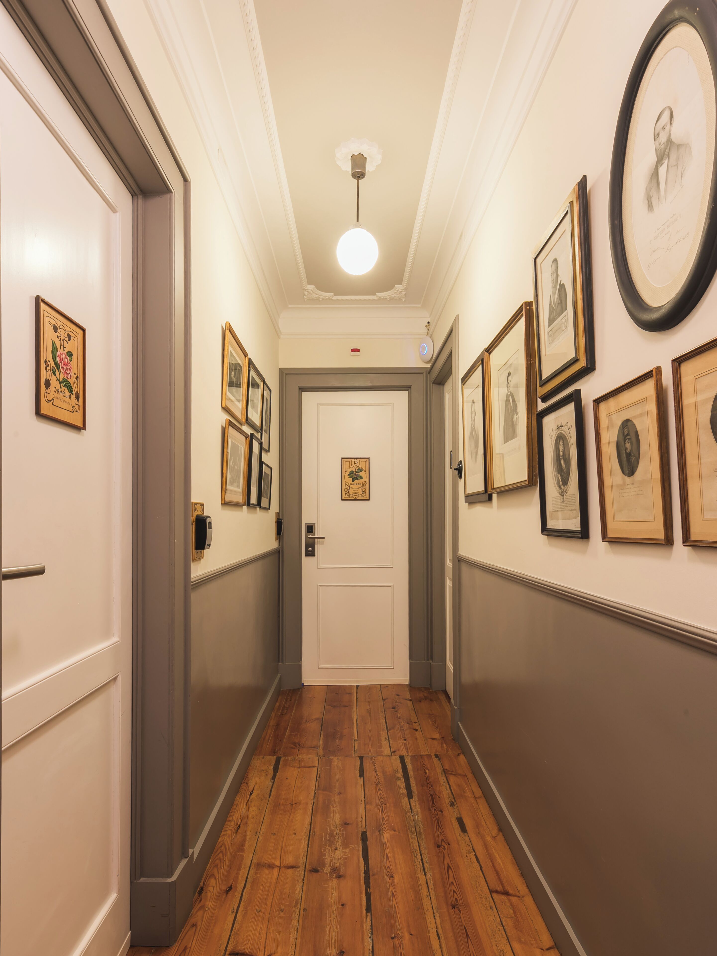 A hallway with framed portraits on the walls, a white door at the end, and wooden flooring.