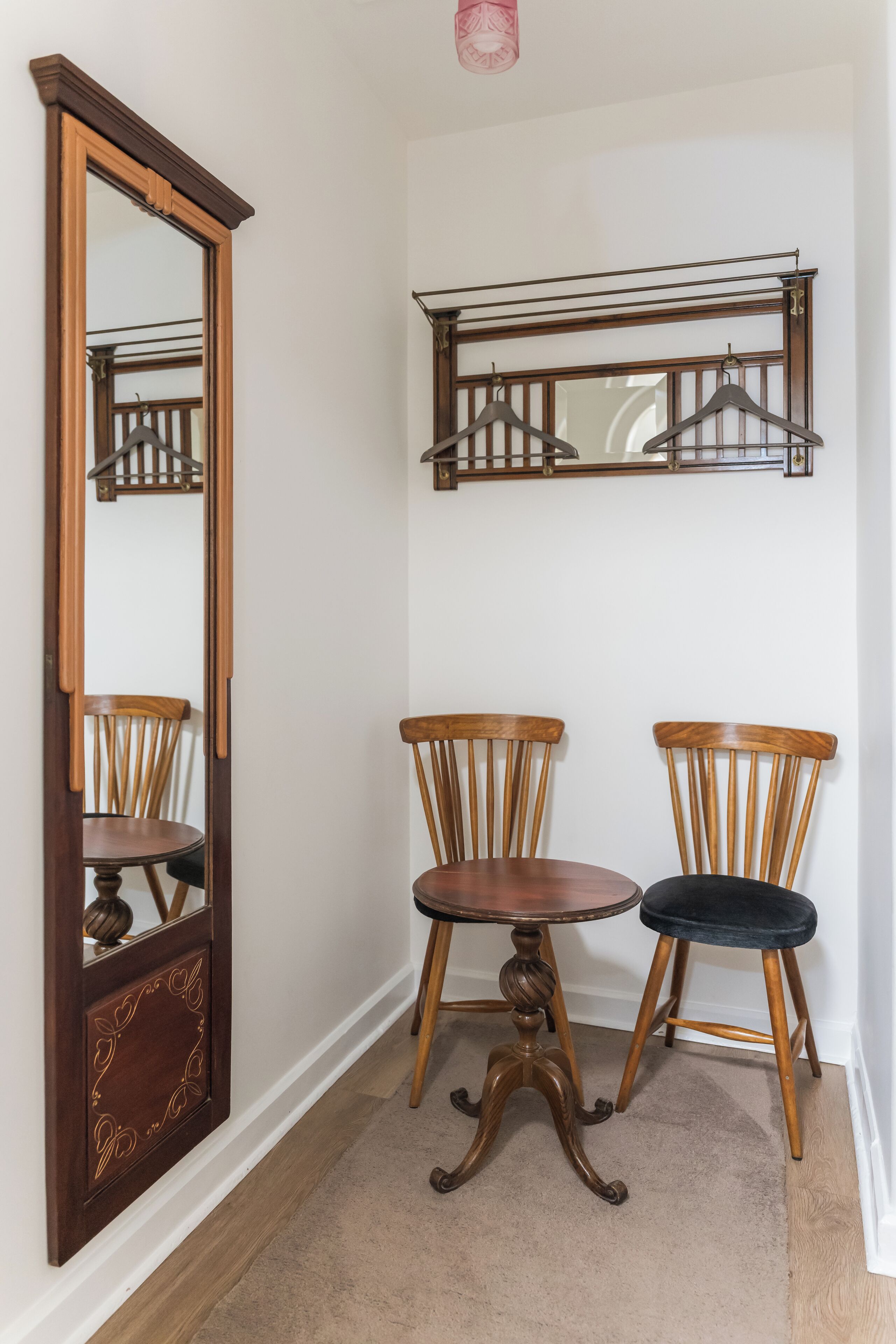 A hallway with a wooden floor, a mirror, two wooden chairs, and a wall-mounted coat rack.