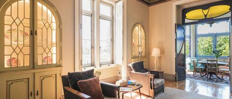 Una habitación con piso de madera, un sofá, una silla, una mesa y una ventana que da a zonas verdes.