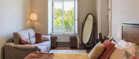 A bedroom with a bed, a sofa, a chair, a lamp, a mirror, and a window with a view of trees.
