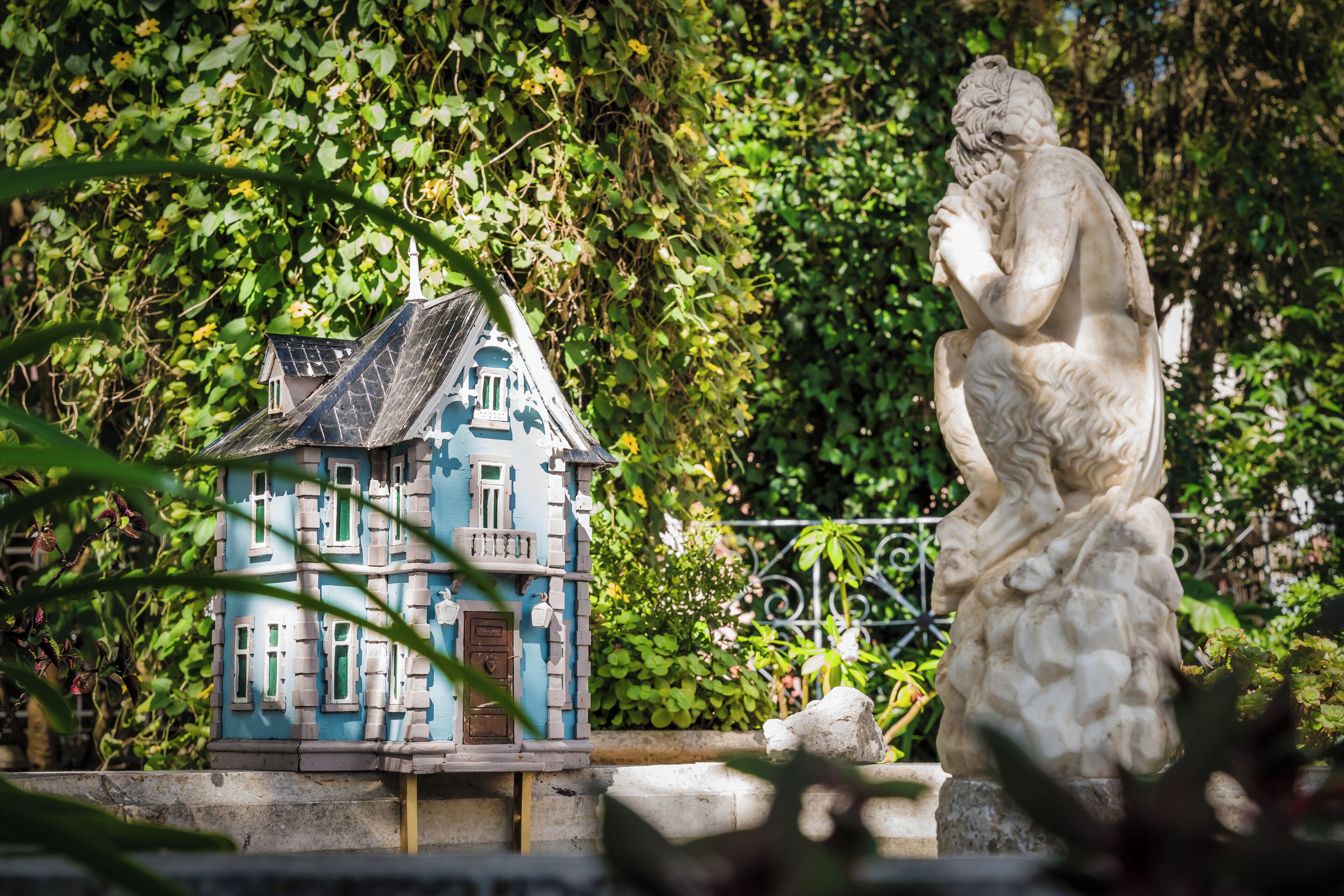 A statue of a seated figure with a contemplative pose, a miniature house model with a blue facade and green windows, and lush greenery in the background.