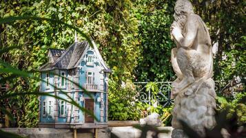 A statue of a seated figure with a contemplative pose, a miniature house model with a blue facade and green windows, and lush greenery in the background.