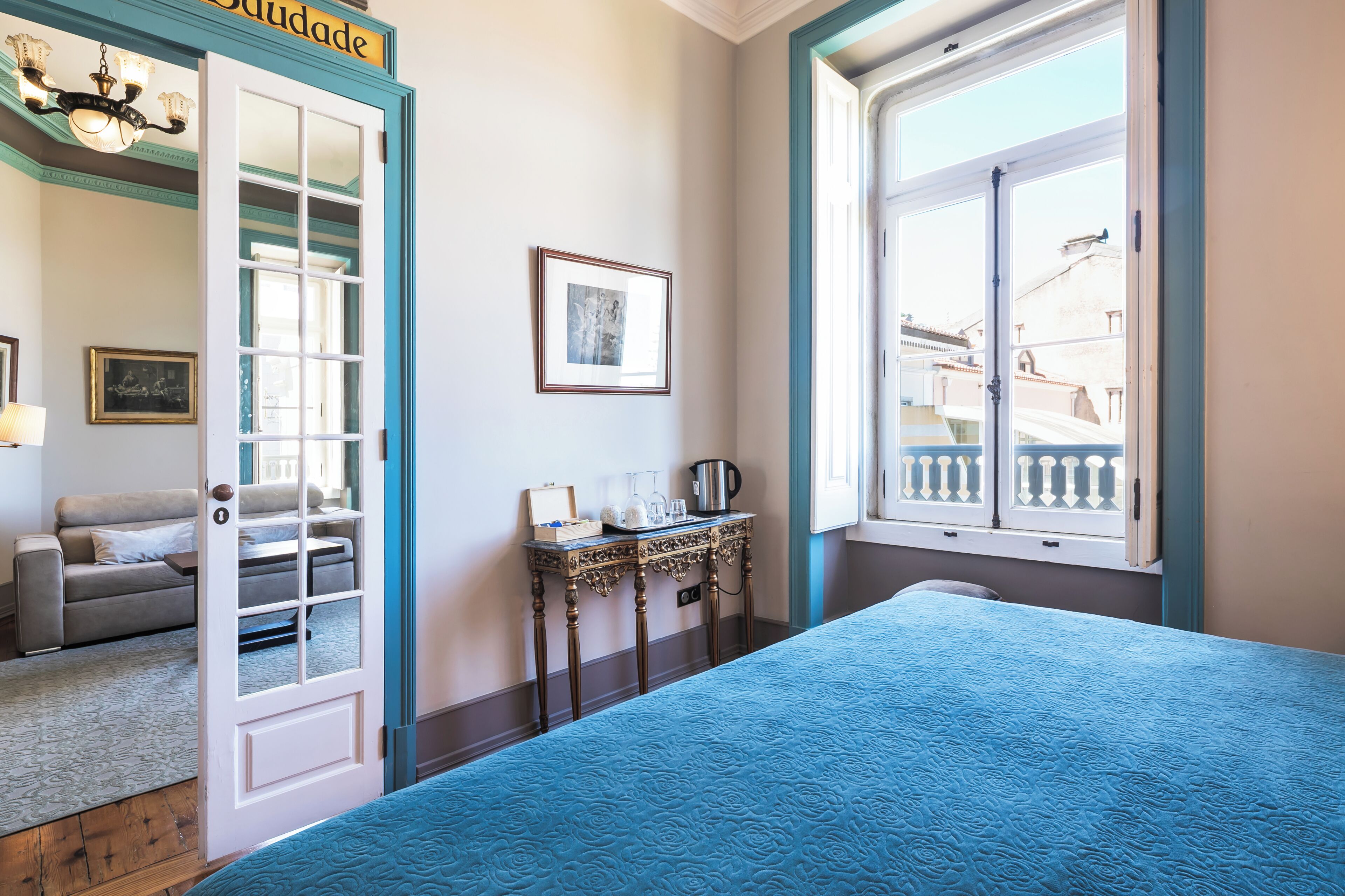 A room with a bed, a table with a kettle, a chair, and a door leading to another room.