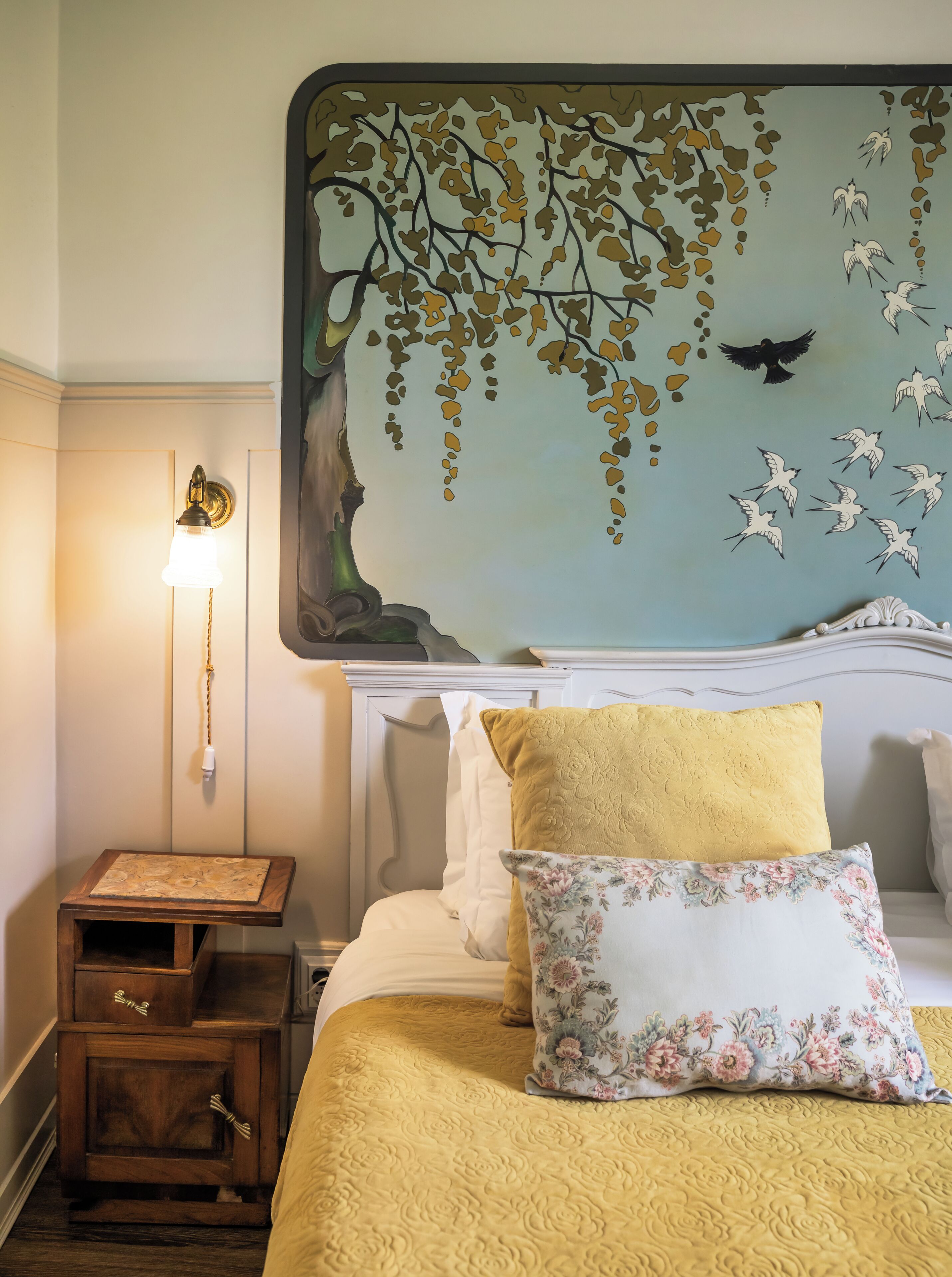 A bedroom with a bed, a nightstand, a wall-mounted lamp, and a wall mural.