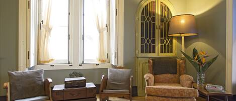 A room with a wooden floor, a window with curtains, a lamp, a chair, a sofa, a table with a vase, and a cabinet.