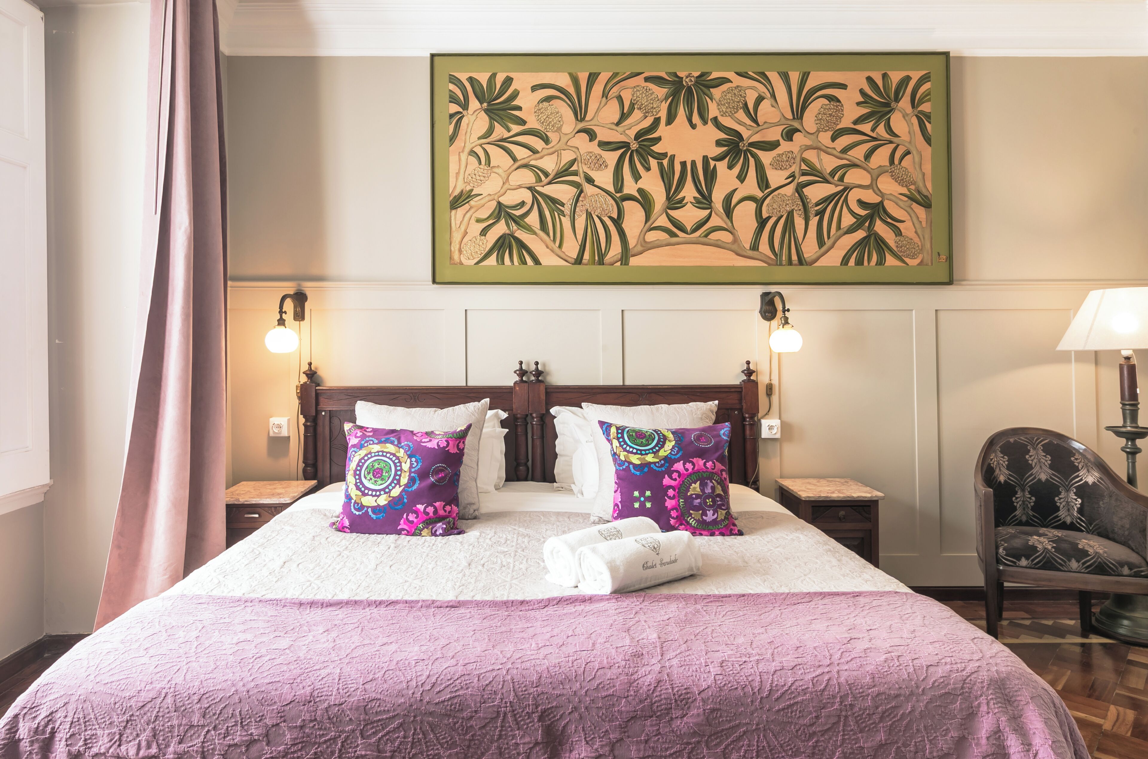 Habitación de hotel con una cama grande, dos mesitas de noche, una silla, un cuadro y una ventana con cortinas.