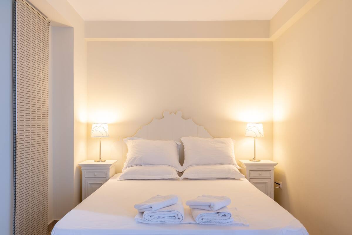 A neatly made bed with two pillows and two folded towels on the bed. There are two bedside tables with lamps on either side of the bed.