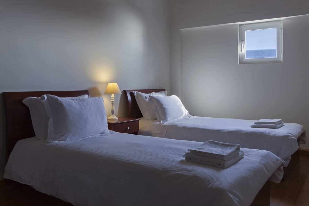 A hotel room with two beds, a nightstand with a lamp, and a window with a view of the sea.